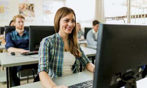 Joven hermosa chica trabajando en una computadora en un aula con sus compañeros de clase en el fondo